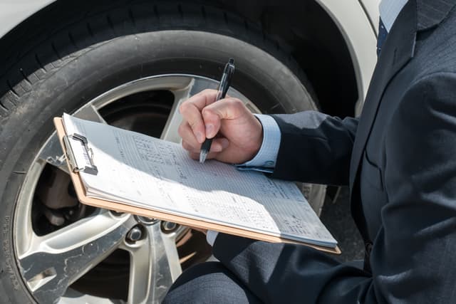 Insurance adjuster or lawyer in a suit taking notes on a clipboard while inspecting a car tire.